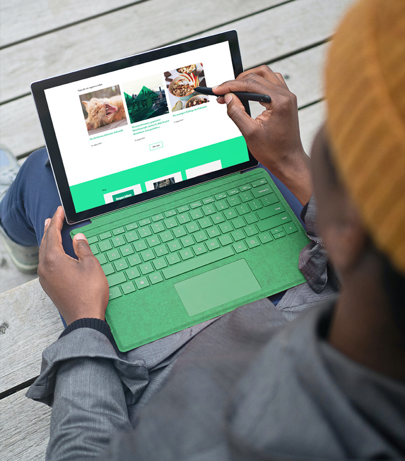 Mensch mit einem Tablet mit grüner Tastatur auf dem Schoß und Stift für Touchscreen in der Hand scrollt durch die vwievegan.de Website.