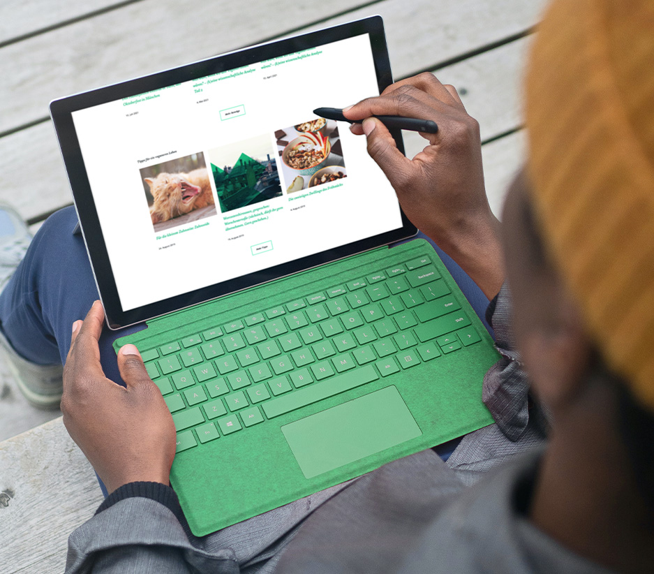 Mensch mit einem Tablet mit grüner Tastatur auf dem Schoß und Stift für Touchscreen in der Hand scrollt durch die vwievegan.de Website.